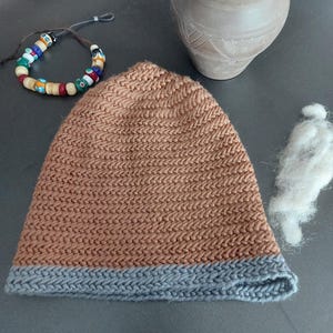 Puede incluir: Un gorro tejido marrón con un borde gris está sobre una superficie oscura. También hay una pulsera de cuentas de colores y un pequeño jarrón de arcilla. Una pequeña cantidad de fibra blanca está al lado del sombrero.