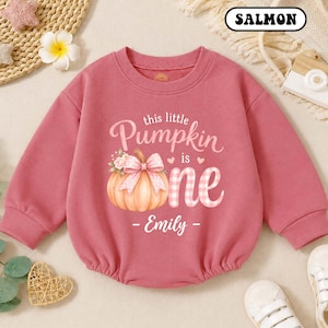 May include: A salmon-colored long-sleeve sweatshirt featuring a graphic of a pumpkin with a pink bow and flowers. The text reads this little Pumpkin is one and Emily. The design includes small hearts and a pink gingham pattern on the word one.