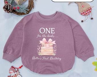 May include: Mauve taupe baby bodysuit with long sleeves. The front features the text "ONE for the books..." above a stack of books tied with a pink bow. The text "Billie's First Birthday" is below the books. The bodysuit is surrounded by holiday decorations.