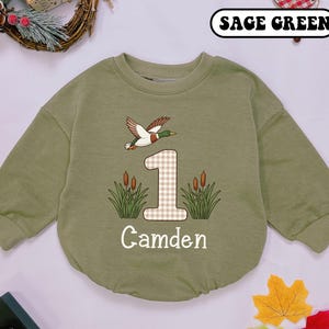 May include: Sage green baby bodysuit with long sleeves. The bodysuit features a flying duck, the number 1 in a plaid pattern, cattails, and the name "Camden" in white. The text "SAGE GREEN" is at the top.