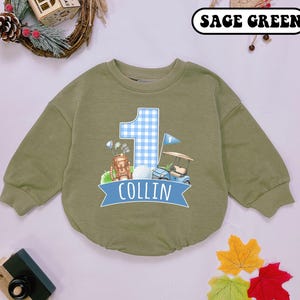 May include: Sage green baby romper with a golf-themed design. Features a large number 1 in blue gingham, a golf cart, golf bag, and a golf ball. The name "COLLIN" is printed on a blue banner.