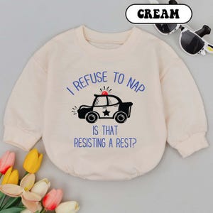 May include: Cream-colored baby romper with long sleeves. The front features a black and white police car graphic and the text "I REFUSE TO NAP IS THAT RESISTING A REST?" A pair of sunglasses is visible in the background.