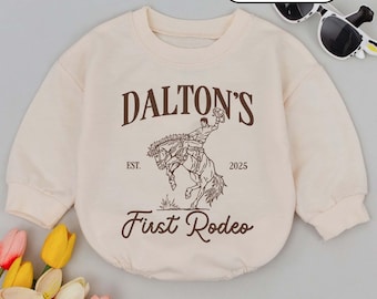 May include: Cream-colored baby romper with a brown graphic of a rodeo rider on a bucking horse. The text reads "DALTON'S" above the image, with "EST. 2025" and "First Rodeo" below. The word "CREAM" is in a white oval.
