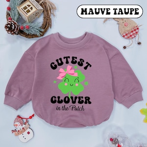 May include: Mauve taupe baby sweatshirt with the text "Cutest Clover in the Patch" and a green clover graphic with a pink bow. The sweatshirt has long sleeves and a round neckline. The background includes holiday decorations.