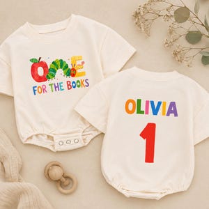 May include: Two cream-colored short-sleeve baby bodysuits. One features a red apple, a green caterpillar, and the text "ONE FOR THE BOOKS". The other displays the name "OLIVIA" in multicolored letters above a large red number "1" on the back.