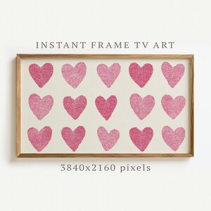 May include: A framed print with fifteen pink hearts on a cream background. The wooden frame surrounds the artwork, with the text "INSTANT FRAME TV ART" above and "3840x2160 pixels" below. The hearts are arranged in three rows.