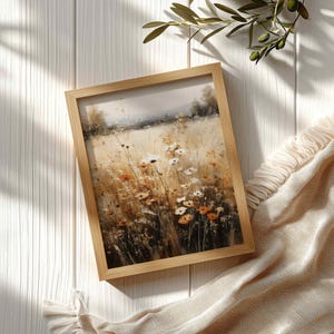 May include: A framed wall art print featuring a painterly landscape of a field filled with wildflowers in shades of white, orange, and gold. The artwork is encased in a light-toned wooden frame, showcasing a soft, hazy horizon line with trees.