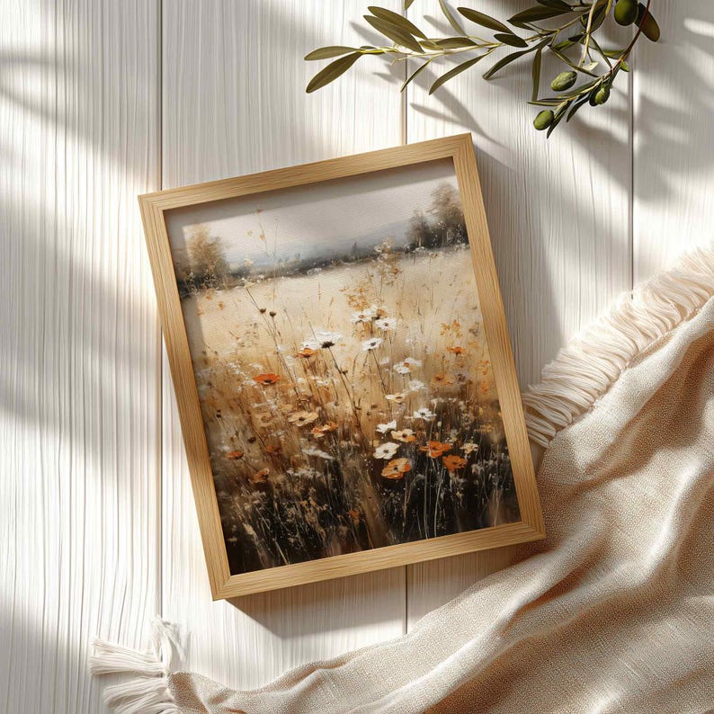 May include: A framed wall art print featuring a painterly landscape of a field filled with wildflowers in shades of white, orange, and gold. The artwork is encased in a light-toned wooden frame, showcasing a soft, hazy horizon line with trees.