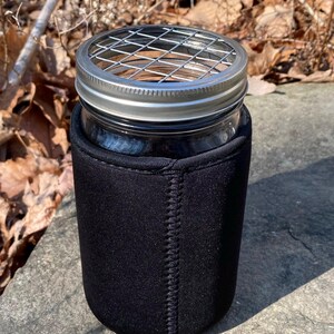 May include: A black neoprene sleeve for a mason jar with a metal wire mesh lid. The jar is sitting on a gray stone surface.