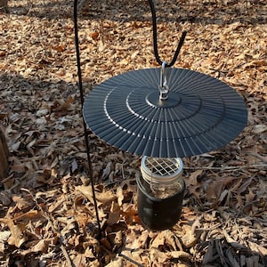 May include: A black metal feeder with a round, black, metal lid and a glass jar hanging from it. The feeder is attached to a black metal stand with a hook at the top. The feeder is sitting on a bed of brown leaves.