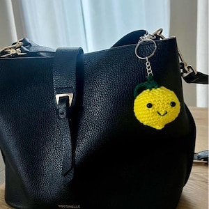 May include: A black leather handbag with a small, yellow crocheted lemon keychain attached. The keychain has a green stem and a smiling face. The handbag has a silver-colored clasp and the brand name "COCCINELLE" is visible.