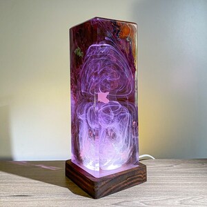 May include: A rectangular resin lamp with a dark wood base. The lamp's interior features swirling purple and white patterns, with hints of red and orange. The lamp is lit, creating a soft glow.