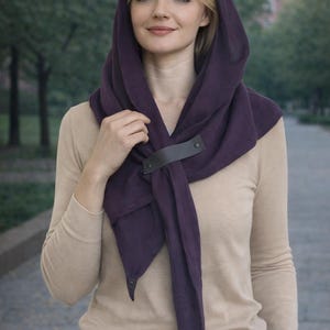 Könnte beinhalten: Ein Kapuzenschal in tiefem Lila, befestigt mit einem dunklen Lederriemen. Der Schal ist über den Schultern drapiert und wird mit einem langärmligen Oberteil in neutralen Tönen und einer grauen Hose getragen. Ein modisches Accessoire.