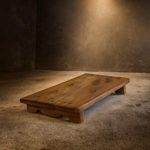 Puede incluir: Un taburete de madera rectangular con un acabado natural. El taburete tiene un perfil bajo, con una parte superior plana y cuatro patas cortas. La veta de la madera es visible y el taburete parece hecho a mano. Las dimensiones son de aproximadamente 61 cm de largo, 30 cm de ancho y 10 cm de alto.