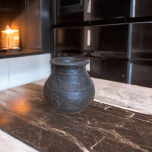 Może przedstawiać: Ciemnoszary, teksturowany ceramiczny wazon o zaokrąglonym korpusie i lekko rozszerzonym brzegu. Wazon stoi na marmurowym blacie, z rozmytym tłem w postaci kuchni.
