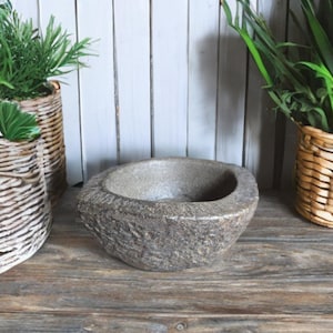 Puede incluir: Un cuenco de piedra gris texturizada con forma ovalada se encuentra sobre una superficie de madera. El cuenco tiene un exterior rugoso y un interior liso. Cestas de mimbre con plantas verdes en el fondo.