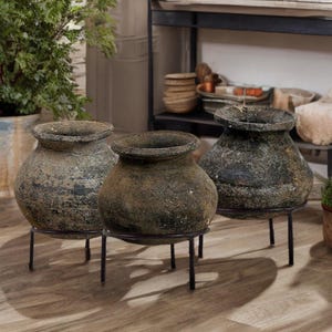 Op de afbeelding: Drie verweerde, getextureerde potten met brede randen en donkere, gevlekte kleuring. Elke pot staat op een zwarte metalen standaard met drie poten. De potten hebben een rustieke, verouderde uitstraling.