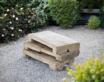 May include: A stack of two weathered, light-brown wooden benches, each with a rectangular top and four short legs. The benches are placed on a bed of small gray stones, with green foliage and pink flowers in the background.
