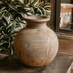 Può includere: Un grande vaso rotondo marrone con una superficie strutturata. Il vaso ha un bordo ampio e leggermente svasato e un collo stretto. La venatura del legno è visibile e il vaso sembra essere fatto di un materiale naturale. Il vaso è un oggetto decorativo.