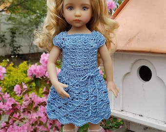 May include: A doll with blonde hair wearing a blue crocheted dress and white socks with black shoes. The doll is standing on a wooden surface in front of a birdhouse.