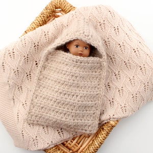 May include: A beige crocheted baby swaddle with a hood, wrapped around a doll with brown eyes and dark hair. The swaddle is lying in a wicker basket lined with a beige crocheted blanket.