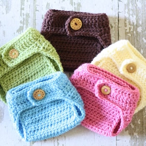 May include: A collection of five handmade crochet baby diapers in various colors: light green, brown, light blue, pink, and cream. Each diaper features a wooden button closure. The diapers are arranged in a circular pattern.