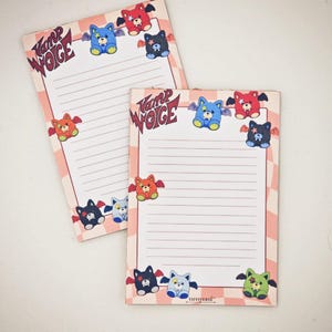 Op de afbeelding: Twee gelijnde notitieblokken met een roze en witte geblokte rand. Elk notitieblok heeft bovenaan de tekst "Vamp Woge" en is versierd met kleurrijke cartoon vleermuisfiguren. De vleermuizen zijn in verschillende kleuren, waaronder blauw, rood en zwart.