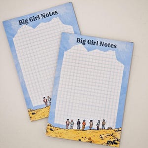 Könnte beinhalten: Zwei Notizblöcke "Big Girl Notes" mit blauem Himmelrand und kariertem Muster. Die Oberseite des Notizblocks hat ein bergähnliches Design. Die Unterseite des Notizblocks zeigt eine gelbe Wüstenlandschaft mit kleinen Figuren. Der Text "Big Girl Notes" ist oben aufgedruckt.