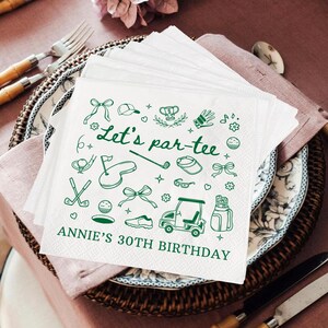 May include: White paper napkins with green golf-themed illustrations and text. The napkins feature golf clubs, golf balls, and a golf cart. The text reads "Let's par-tee" and "Annie's 30th Birthday". The napkins are stacked on a decorative plate.