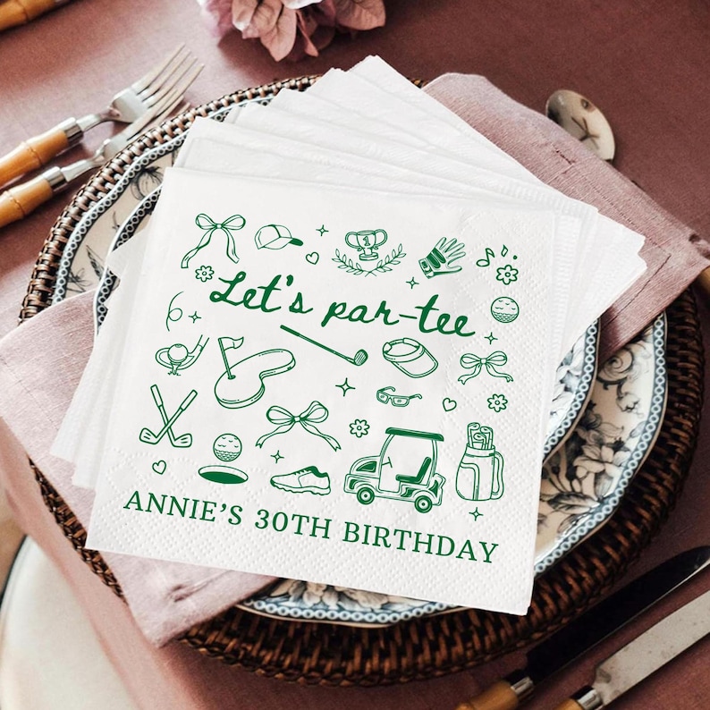 May include: White paper napkins with green golf-themed illustrations and text. The napkins feature golf clubs, golf balls, and a golf cart. The text reads "Let's par-tee" and "Annie's 30th Birthday". The napkins are stacked on a decorative plate.