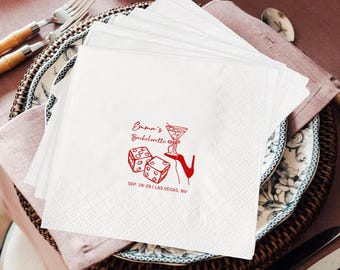 Op de afbeelding: Witte papieren servetten met een rode afbeelding van dobbelstenen, een cocktail en een hoge hak. De tekst "Emma's Bachelorette" staat boven de afbeelding, met "SEP. 28-29 | LAS VEGAS, NV" eronder. Servetten zijn op een decoratieve plaat gestapeld.