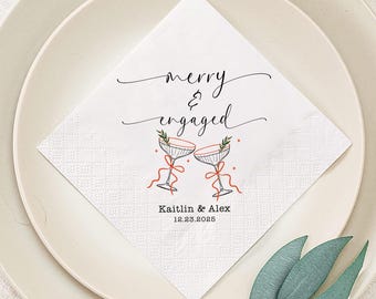 May include: White square paper napkin with the words "merry & engaged" in elegant script. Two champagne glasses with red ribbons and greenery are illustrated below the text. The names "Kaitlin & Alex" and the date "12.23.2025" are printed below the glasses.