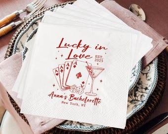 Op de afbeelding: Witte papieren servetten met een rood ontwerp met speelkaarten, dobbelstenen en een martiniglas. De tekst luidt "Lucky in Love" en "Anna's Bachelorette, New York, NY". De servetten zijn op een decoratief bord gestapeld.