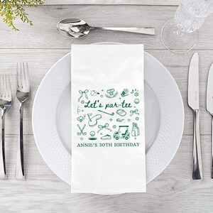 May include: A white paper napkin with green golf-themed illustrations and text. The text reads "Let's par-tee" and "Annie's 30th Birthday". The napkin is placed on a white plate, with silverware and a glass visible.