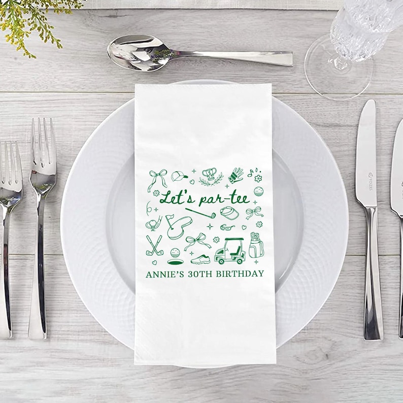 May include: A white paper napkin with green golf-themed illustrations and text. The text reads "Let's par-tee" and "Annie's 30th Birthday". The napkin is placed on a white plate, with silverware and a glass visible.