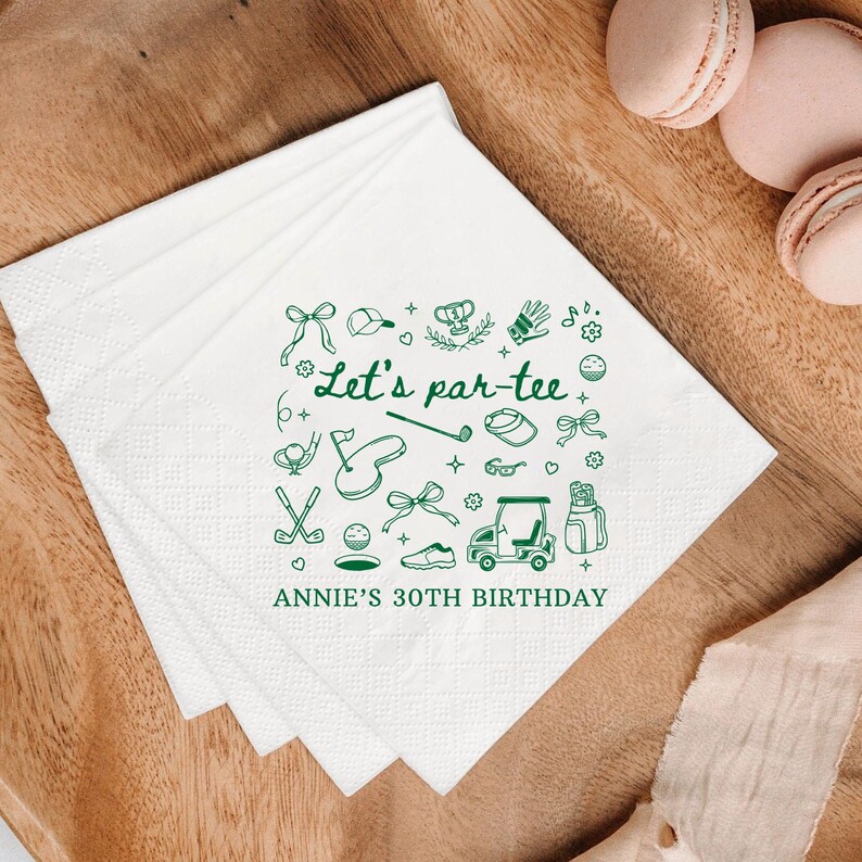 May include: White paper napkins with a green golf-themed design. The napkins feature the text "Let's par-tee" and "Annie's 30th Birthday" along with golf-related illustrations. The napkins are stacked on a wooden surface.