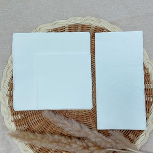 May include: A stack of white paper napkins, folded and arranged on a woven wicker placemat. A sprig of dried, feathery pampas grass is placed near the napkins. The napkins are a bright white color.
