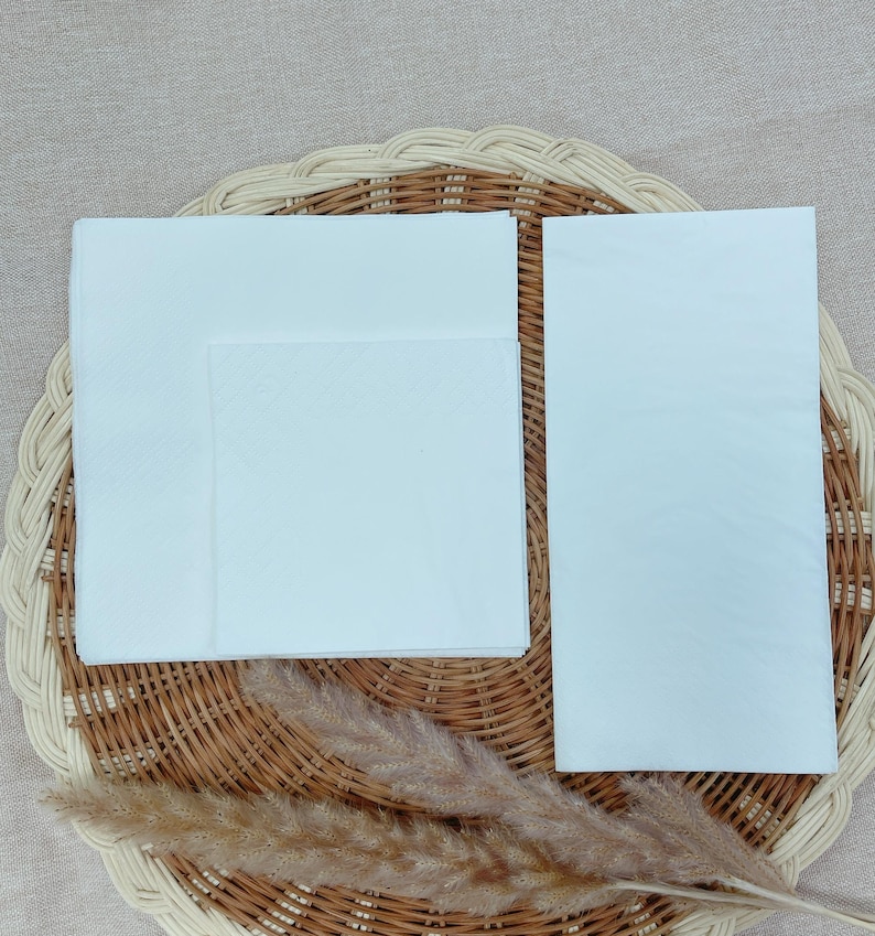 May include: A stack of white paper napkins, folded and arranged on a woven wicker placemat. A sprig of dried, feathery pampas grass is placed near the napkins. The napkins are a bright white color.