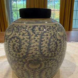 Op de afbeelding: Een ronde keramische gemberpot met een witte basis, blauwe handgeschilderde bloemrankpatronen en een geometrische rand aan de bovenzijde. Het vat is afgesloten met een massief zwart, rond deksel dat naadloos op de opening aansluit.