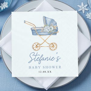 May include: A baby shower napkin with a watercolor illustration of a blue baby carriage, adorned with flowers. The text "Stefanie's BABY SHOWER" is written in elegant script. The napkin is set on a white plate with silverware and a blue tablecloth.