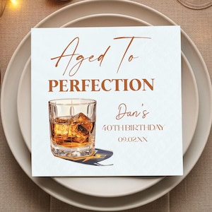 May include: White square napkin with the text "Aged To Perfection" and "Dan's 40th Birthday 09.02.XX" in copper-colored script. A watercolor illustration of a whiskey glass with ice cubes is featured below the text. The napkin is on a white plate.