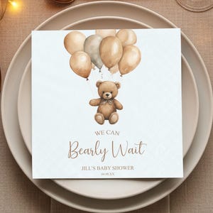 May include: A baby shower napkin featuring a watercolor illustration of a teddy bear holding balloons. The text "We Can Bearly Wait" is displayed below the bear, with the baby shower details at the bottom. The napkin is placed on a white plate with gold cutlery.