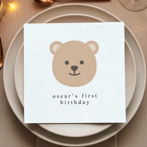 May include: A white square napkin with a light brown teddy bear face and the text "oscar's first birthday" in black. The napkin is on a white plate, with gold-colored cutlery and candles in the background.