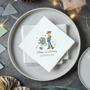 Puede incluir: Servilletas de papel cuadradas blancas con una ilustraci&oacute;n de dibujos animados de Buzz Lightyear y Woody de Toy Story. El texto "Happy 2nd Birthday CAMERON" est&aacute; impreso debajo. Las servilletas est&aacute;n apiladas en un plato blanco.