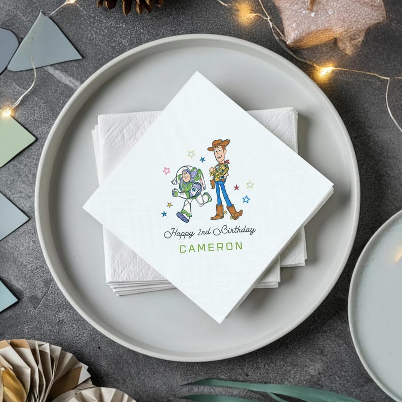Puede incluir: Servilletas de papel cuadradas blancas con una ilustraci&oacute;n de dibujos animados de Buzz Lightyear y Woody de Toy Story. El texto "Happy 2nd Birthday CAMERON" est&aacute; impreso debajo. Las servilletas est&aacute;n apiladas en un plato blanco.