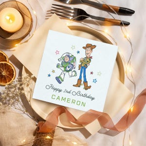 Puede incluir: Una tarjeta de cumplea&ntilde;os blanca con ilustraciones de Buzz Lightyear y Woody, rodeada de estrellas de colores. La tarjeta dice "Happy 2nd Birthday CAMERON" en escritura negra y verde. La tarjeta est&aacute; sobre un plato con una cinta y cubiertos.