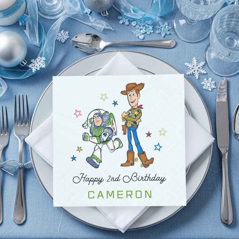 Puede incluir: Una servilleta cuadrada blanca con una ilustraci&oacute;n colorida de Woody y Buzz Lightyear de Toy Story. La servilleta dice "Happy 2nd Birthday CAMERON" en texto verde y negro. La servilleta est&aacute; colocada sobre un plato blanco.