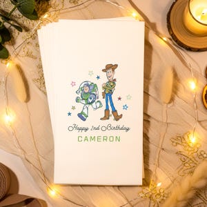 Puede incluir: Un mont&oacute;n de servilletas de papel blanquecino con una colorida ilustraci&oacute;n de Buzz Lightyear y Woody de Toy Story. El texto "Happy 2nd Birthday CAMERON" est&aacute; impreso debajo, con estrellas.