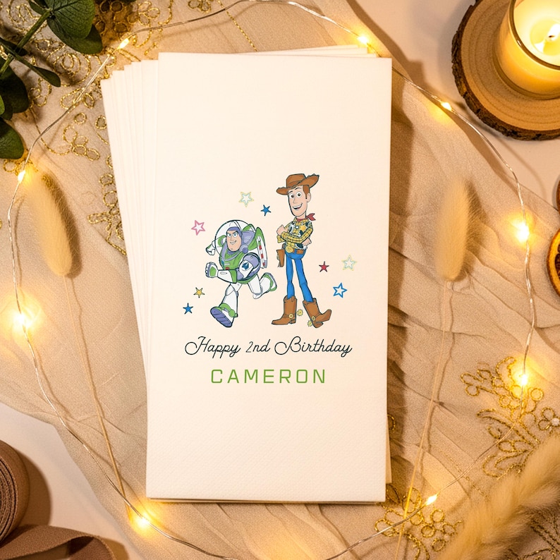 Puede incluir: Un mont&oacute;n de servilletas de papel blanquecino con una colorida ilustraci&oacute;n de Buzz Lightyear y Woody de Toy Story. El texto "Happy 2nd Birthday CAMERON" est&aacute; impreso debajo, con estrellas.