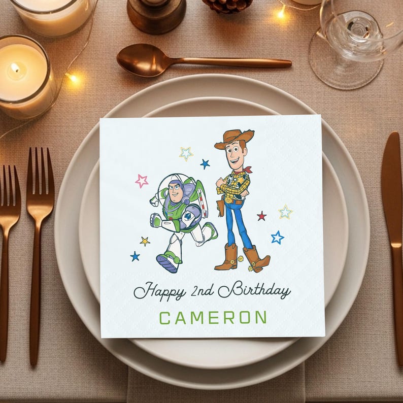 Puede incluir: Una servilleta cuadrada blanca con una ilustraci&oacute;n de Buzz Lightyear y Woody de Toy Story, con el texto "Happy 2nd Birthday CAMERON" en verde y negro. La servilleta est&aacute; sobre un plato blanco, con un tenedor y un cuchillo.